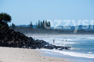 Lennox Head Beach from Seven Mile Beach. With a backdrop of greenery, there are people fishing, swimming, and enjoying the beach.
