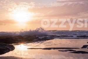 Stunning stock photo of Shelly Beach at sunrise, featuring waves crashing onto rocks and a serene rock pool reflecting the beautiful colors of the sky.
