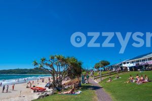 Byron Bay Main Beach and Apex Park, with sunbathers, surf life saving club, and the iconic lighthouse in the background.