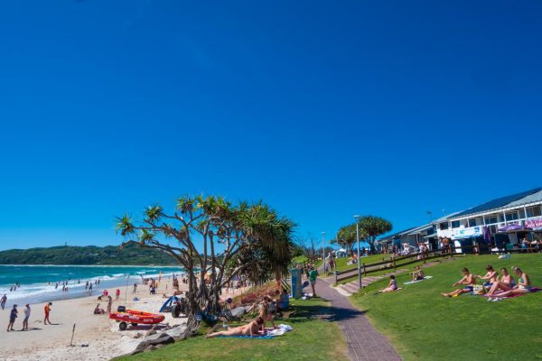Byron Bay Main Beach and Apex Park, with sunbathers, surf life saving club, and the iconic lighthouse in the background.